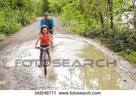 Stock Image - Father and daughter riding bicycles through puddle. Fotosearch