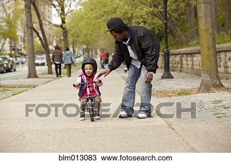 Stock Image - Father helping daughter ride bicycle on sidewalk. Fotosearch
