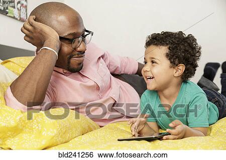 Stock Photograph - Father watching son laying on bed using digital tablet. Fotosearch