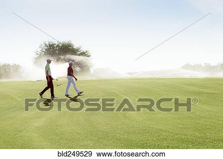 Stock Photo - Friends walking on golf course near sprinklers. Fotosearch