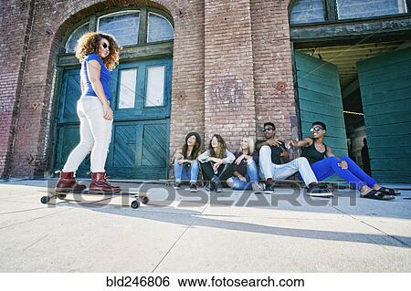 Stock Photograph - Friends watching woman skateboarding on sidewalk. Fotosearch