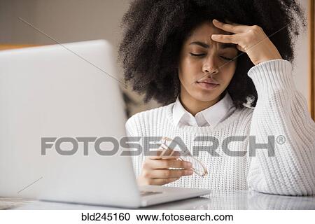 Stock Image - Frustrated African American woman holding eyeglasses near laptop. Fotosearch