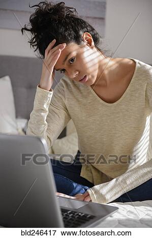 Stock Photo - Frustrated Hispanic woman sitting on bed using laptop. Fotosearch