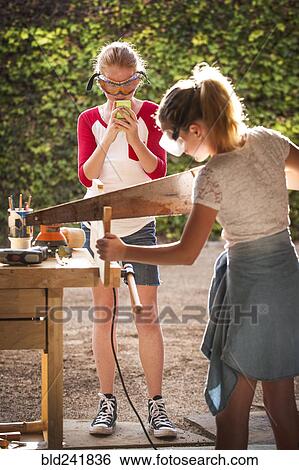Stock Photograph - Girl photographing sister sawing wood in garage. Fotosearch