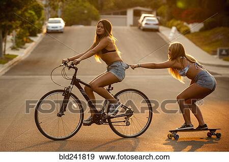 Stock Photo - Girl riding bicycle pulling friend on skateboard. Fotosearch