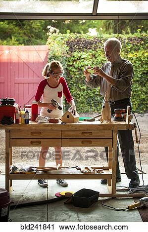 Stock Image - Grandfather photographing granddaughter building birdhouse in garage. Fotosearch