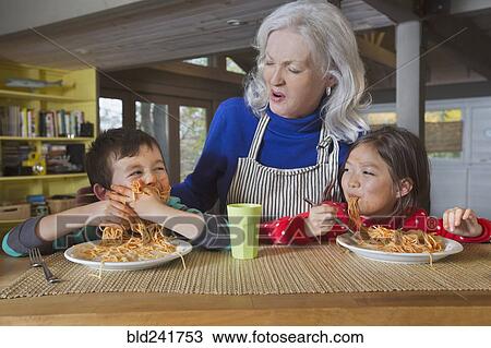 Stock Image - Grandmother watching grandson and granddaughter eat spaghetti. Fotosearch