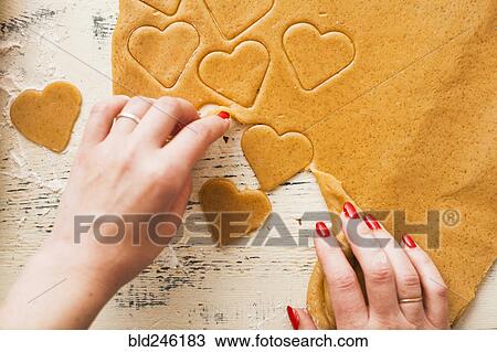 Stock Image - Hands of woman separating heart shapes cut from cookie dough. Fotosearch