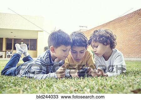 Stock Image - Hispanic boys laying in grass texting on cell phone. Fotosearch