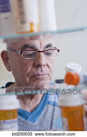 Stock Image - Hispanic man examining pill bottle from medicine cabinet. Fotosearch