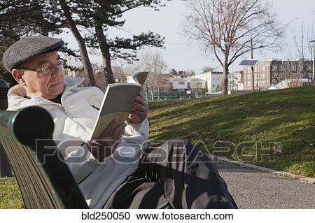 Stock Image - Hispanic man sitting on park bench reading book. Fotosearch