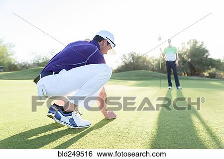 Stock Photograph - Hispanic man watching friend crouching on golf course. Fotosearch