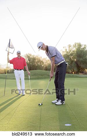 Stock Image - Hispanic man watching friend on golf course. Fotosearch