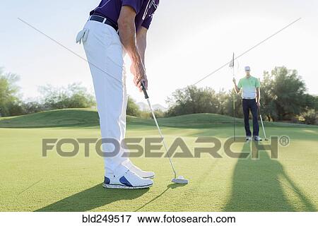 Stock Photo - Hispanic man watching friend putting on golf course. Fotosearch