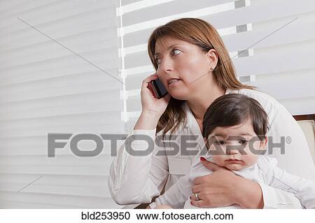 Stock Image - Hispanic mother holding baby boy and talking on cell phone. Fotosearch