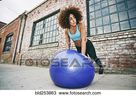 Stock Image - Hispanic woman balancing on fitness ball near brick wall. Fotosearch