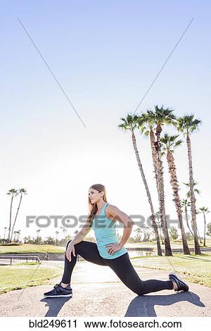 Stock Image - Hispanic woman kneeling and stretching on path. Fotosearch