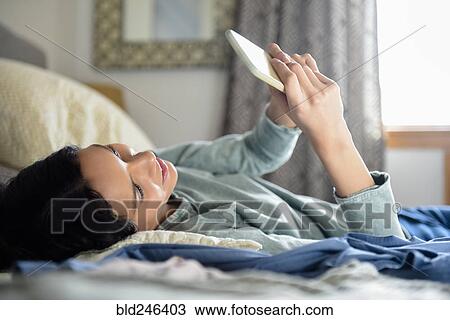 Stock Image - Hispanic woman laying in bed texting on cell phone. Fotosearch