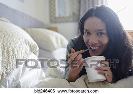 Stock Photograph - Hispanic woman laying on bed eating ice cream from carton. Fotosearch