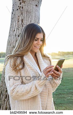 Picture - Hispanic woman leaning on tree texting on cell phone. Fotosearch