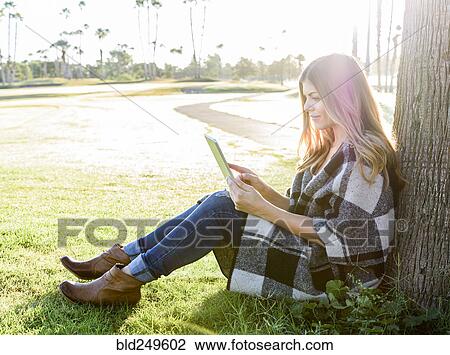 Stock Image - Hispanic woman leaning on tree using digital tablet. Fotosearch