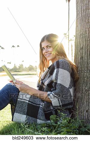Stock Image - Hispanic woman leaning on tree using digital tablet. Fotosearch