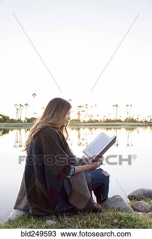 Stock Image - Hispanic woman sitting bear lake reading book. Fotosearch