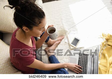Hispanic woman sitting on bed drinking tea and using laptop View Large Photo Image Stock Photo - Hispanic woman sitting on bed drinking tea and using laptop. Fotosearch