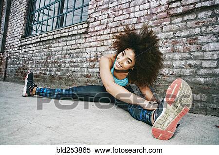 Stock Photography - Hispanic woman sitting on sidewalk stretching legs near brick wall. Fotosearch