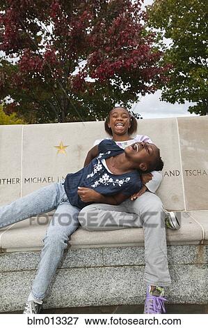 Stock Photo - Laughing Black girls sitting on bench. Fotosearch