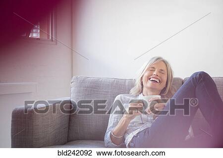 Stock Image - Laughing Caucasian woman sitting on sofa drinking coffee. Fotosearch