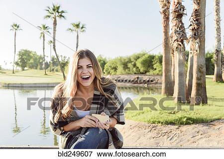 Laughing Hispanic woman holding cell phone near pond View Large Photo Image Stock Photo - Laughing Hispanic woman holding cell phone near pond. Fotosearch