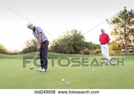 Stock Photo - Man watching friend putting on golf course. Fotosearch