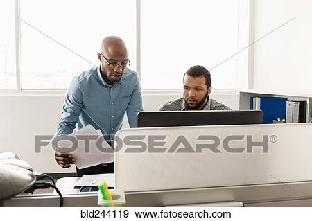 Stock Photo - Men working at computer with paperwork in office. Fotosearch