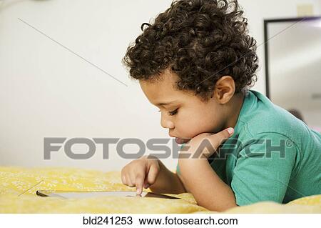 Stock Image - Mixed Race boy laying on bed using digital tablet. Fotosearch