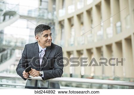 Stock Image - Mixed Race businessman leaning on railing in lobby holding cell phone. Fotosearch