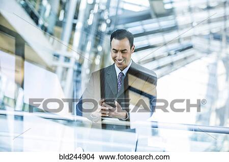 Picture - Mixed Race businessman texting on cell phone leaning on railing in lobby. Fotosearch