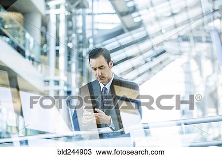 Stock Image - Mixed Race businessman texting on cell phone leaning on railing in lobby. Fotosearch