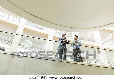 Stock Image - Mixed Race businessmen talking in lobby using digital tablet. Fotosearch