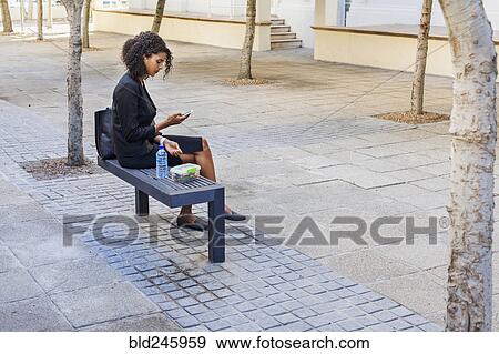 Stock Photo - Mixed Race businesswoman eating lunch on bench texting on cell phone. Fotosearch