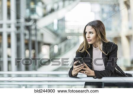 Stock Image - Mixed Race businesswoman texting on cell phone leaning on railing in lobby. Fotosearch