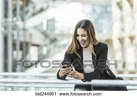 Stock Image - Mixed Race businesswoman texting on cell phone leaning on railing in lobby. Fotosearch
