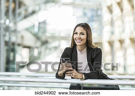 Stock Image - Mixed Race businesswoman texting on cell phone leaning on railing in lobby. Fotosearch