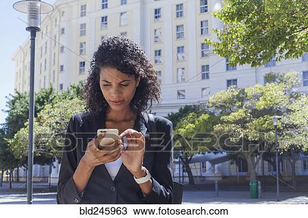 Stock Image - Mixed Race businesswoman texting on cell phone outdoors. Fotosearch