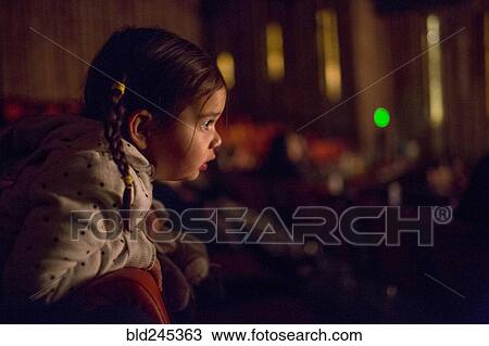 Stock Image - Mixed Race girl leaning on chair watching movie in theater. Fotosearch