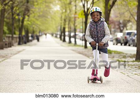 Stock Image - Mixed race girl riding on push scooter on sidewalk. Fotosearch
