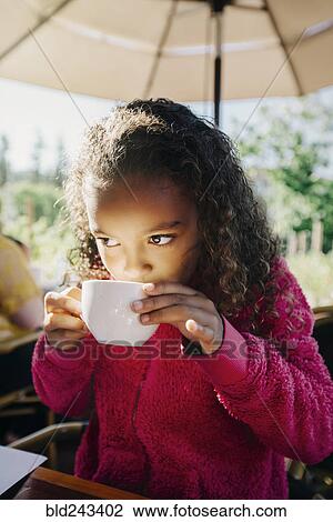 Stock Image - Mixed Race girl sipping from cup in restaurant. Fotosearch