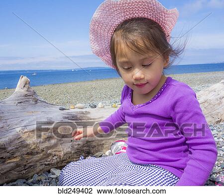 Stock Image - Mixed Race girl sitting on beach near log. Fotosearch