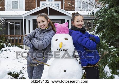 Stock Image - Mixed Race girls posing with snowman wearing pink hat with ears. Fotosearch