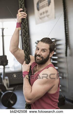 Stock Photo - Mixed Race man climbing rope gymnasium. Fotosearch